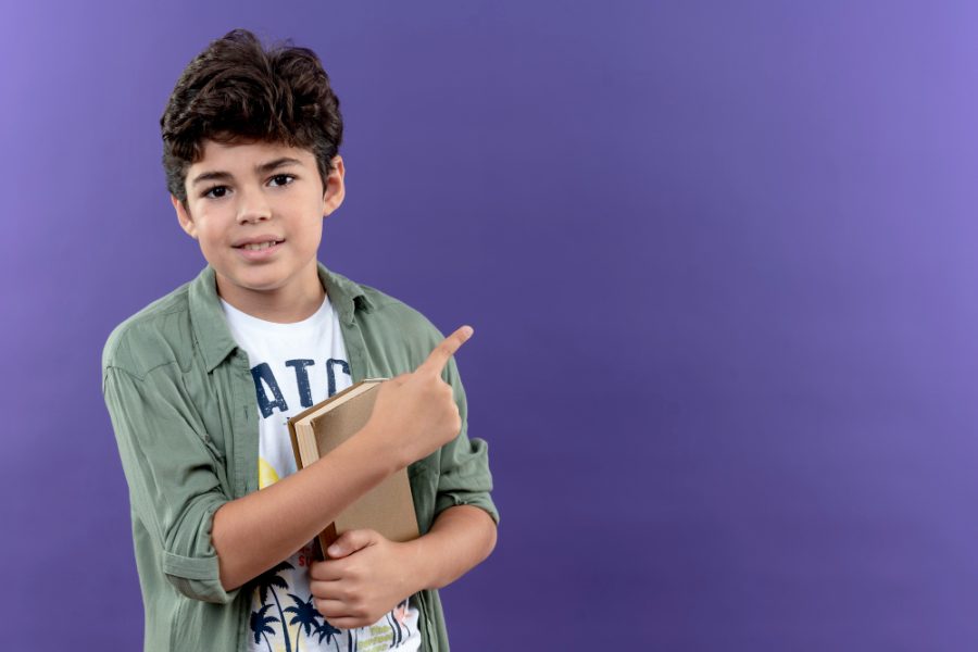 pleased-little-schoolboy-holding-book-points-side-isolated-purple-wall-with-copy-space