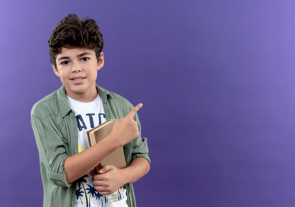 pleased-little-schoolboy-holding-book-points-side-isolated-purple-wall-with-copy-space