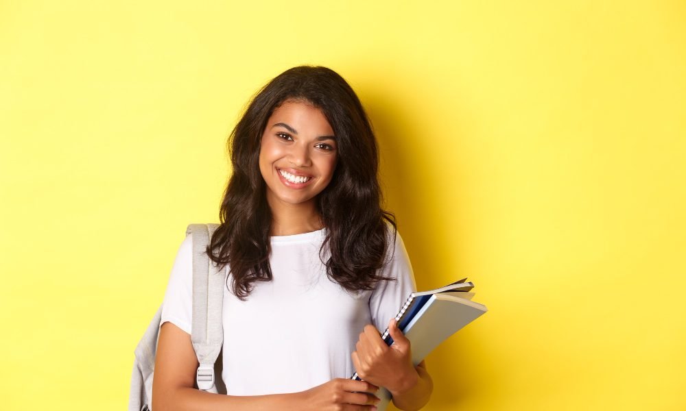 portrait-happy-african-american-female-college-student-holding-notebooks-backpack-smiling-standing-yellow-background