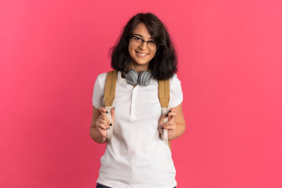 young-smiling-pretty-caucasian-schoolgirl-wearing-glasses-headphones-neck-back-bag-looks-camera-pink-with-copy-space