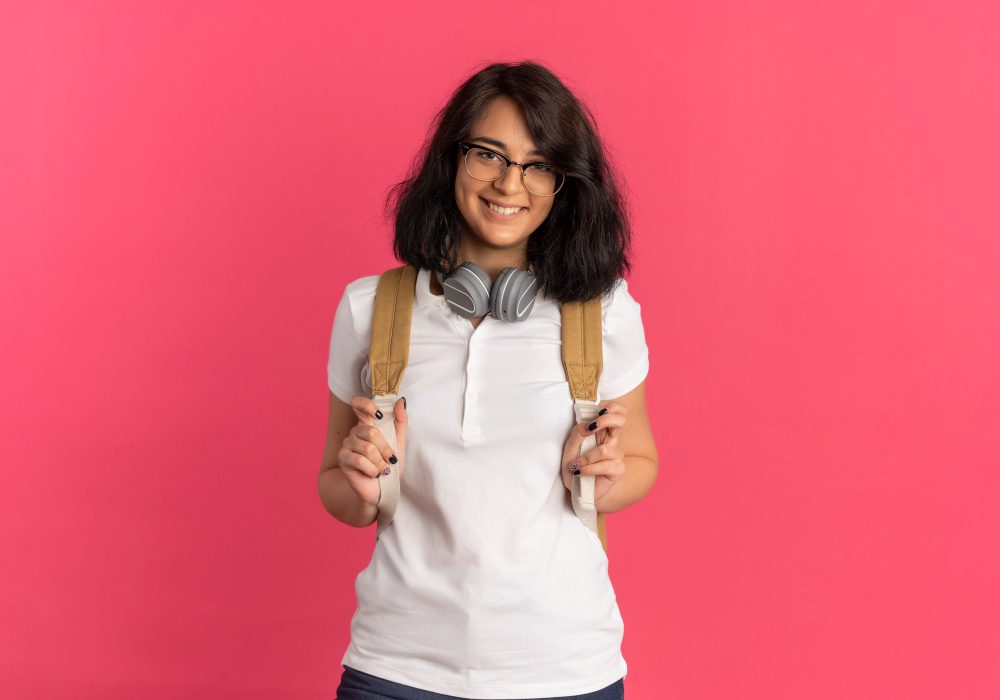 young-smiling-pretty-caucasian-schoolgirl-wearing-glasses-headphones-neck-back-bag-looks-camera-pink-with-copy-space